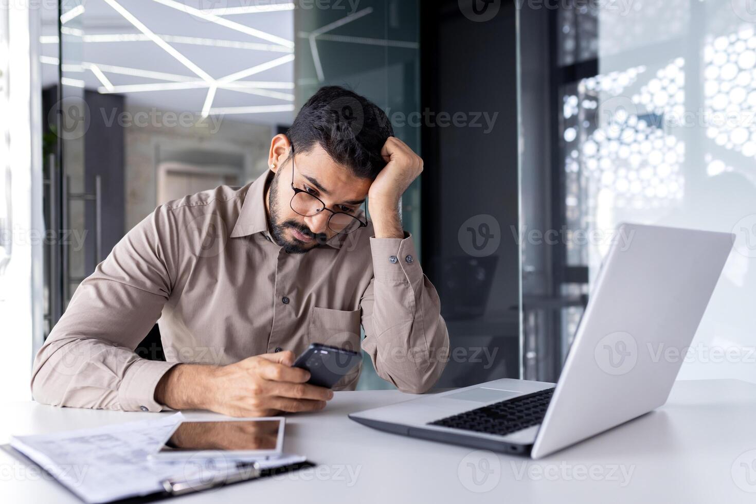 Upset sad man at workplace inside office, businessman uses phone reads bad news received by e-mail, employee disappointed holds smartphone in hands browses internet sites and social networks. photo
