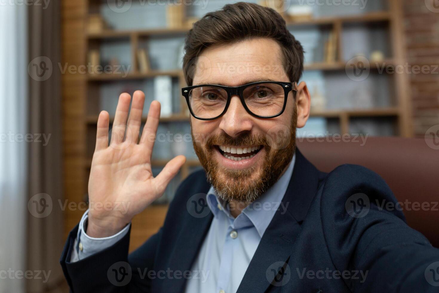 Friendly mature businessman with glasses waving hello, working from a cozy home office with classic decor. photo