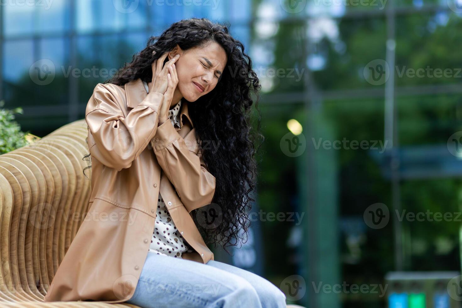 Frustrated woman sitting on modern curved bench outdoors. Disappointment female holding hand to ear and feeling discomfort or pain in that area. Background of building with large glass windows. photo