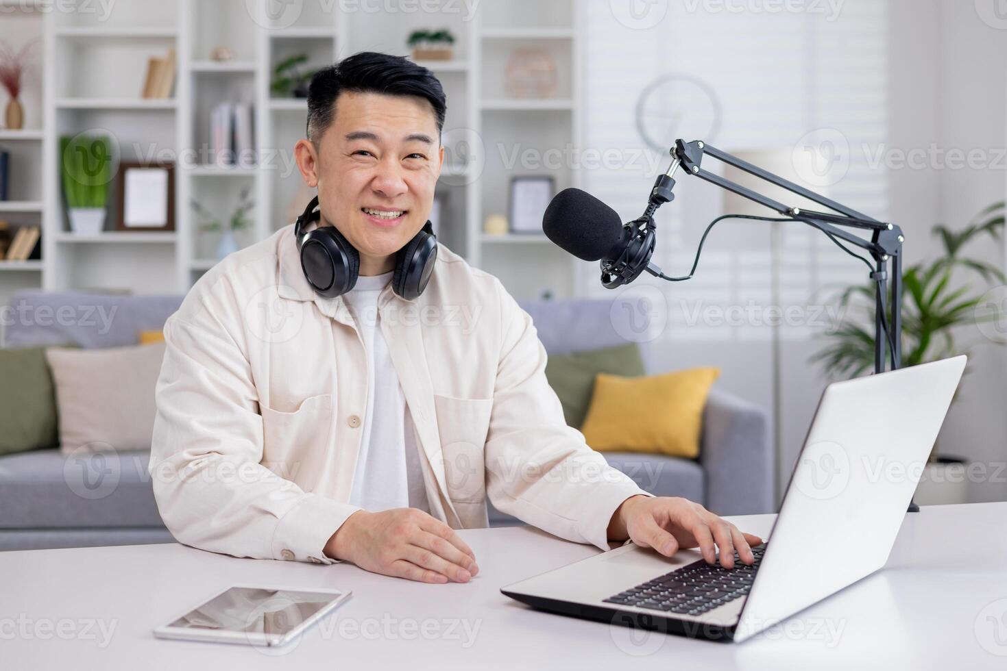 Portrait of blogger at home, Asian man smiling and looking at camera, man sitting at table with laptop in living room, recording audio podcasts and doing online radio. photo