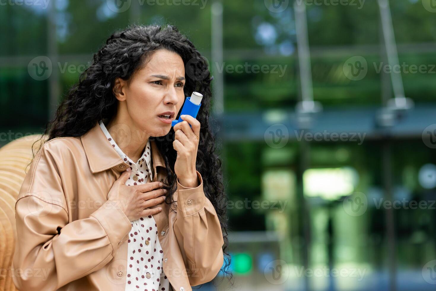 Concerned young Hispanic businesswoman using an asthma inhaler during a break, with an office building in the background. photo