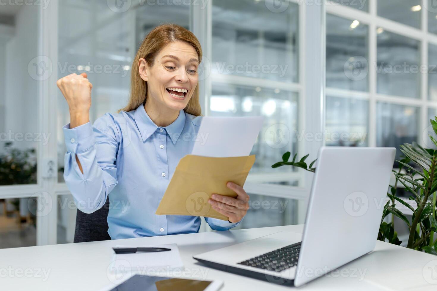 A joyful female office worker celebrates success with an envelope in a modern workspace, evoking feelings of achievement and happiness. photo