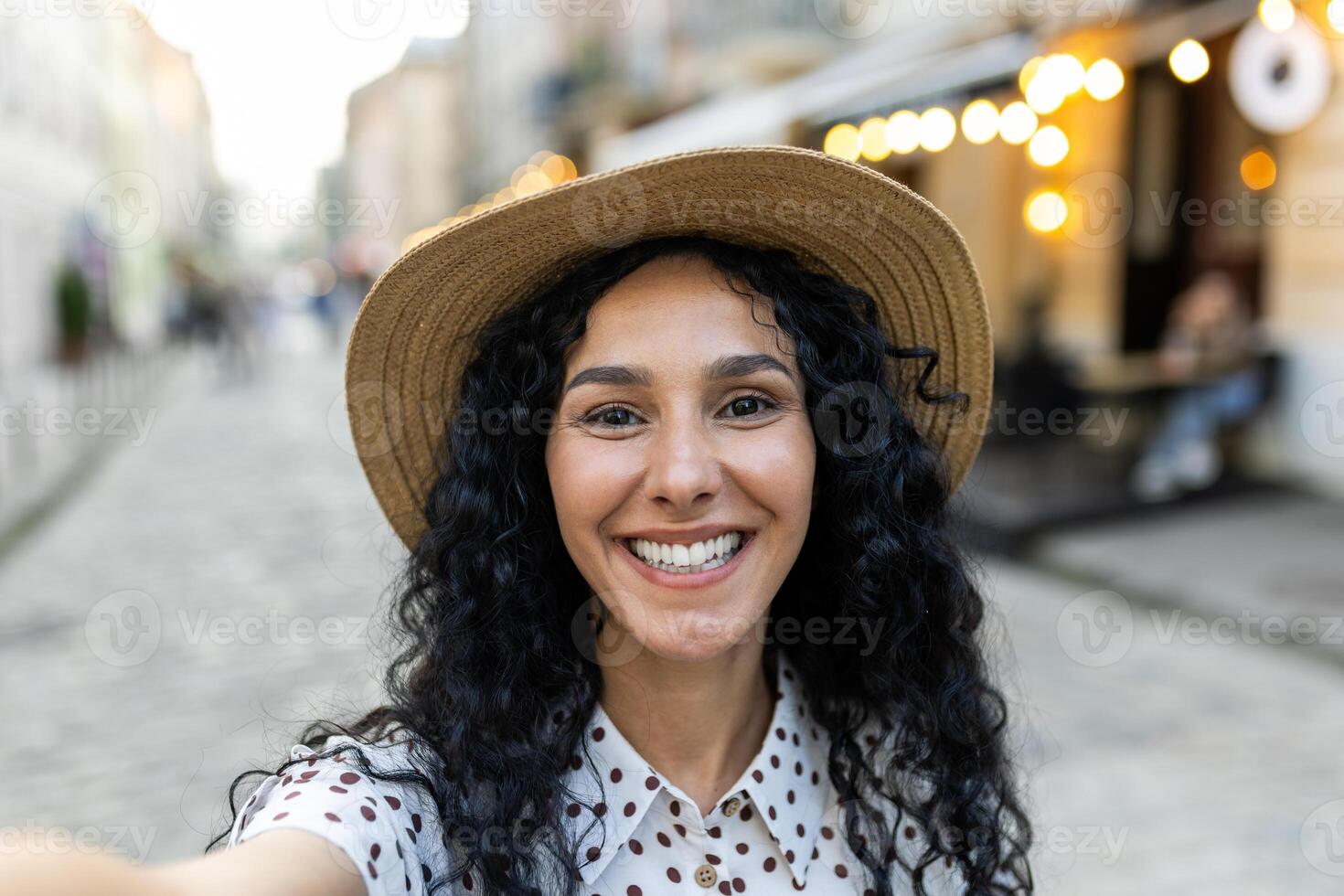 Young beautiful hispanic woman smiling and looking into phone camera, traveler uses smartphone app for remote communication call, walks in evening city, on tourist tour trip. photo