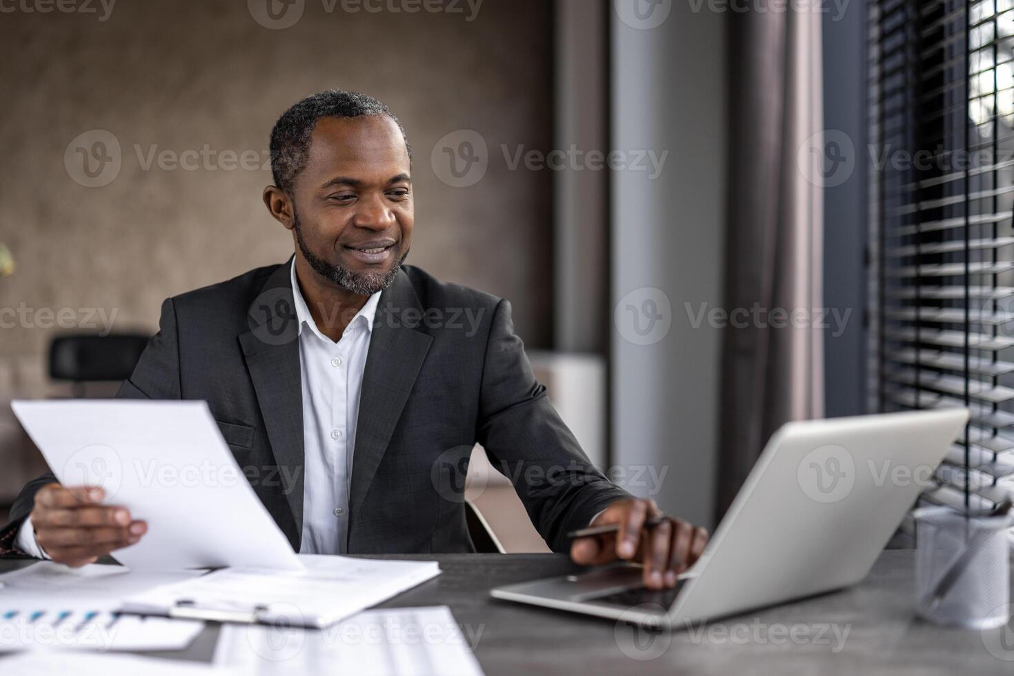 Confident African American businessman in formal attire working on a laptop while reviewing documents at his office desk. photo
