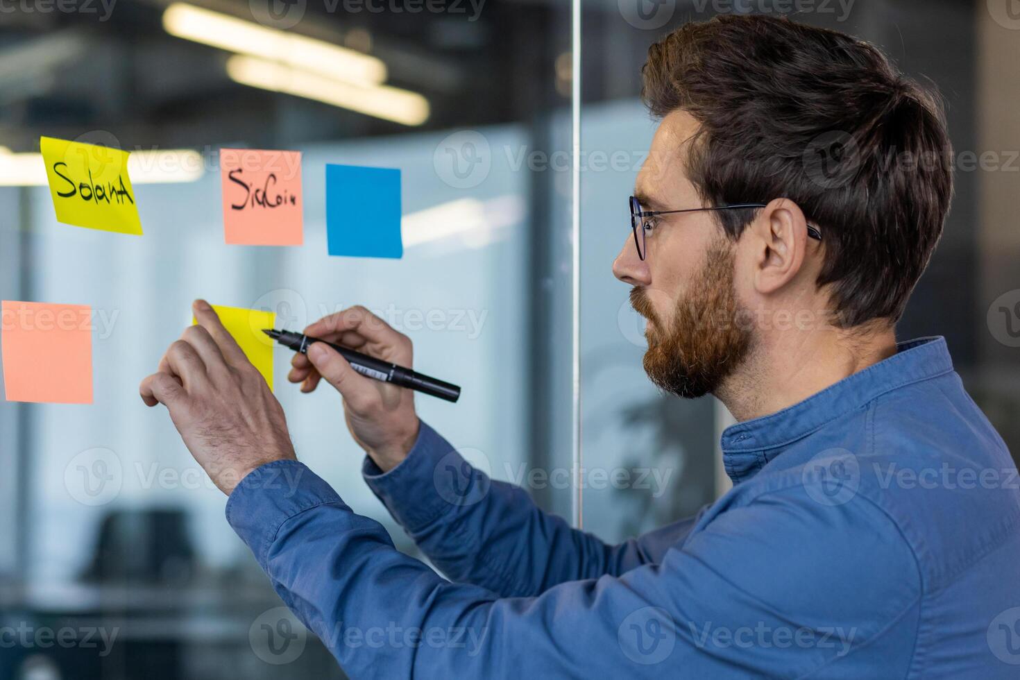 Close-up photo of a young businessman standing in an office and taking notes on a glass board.