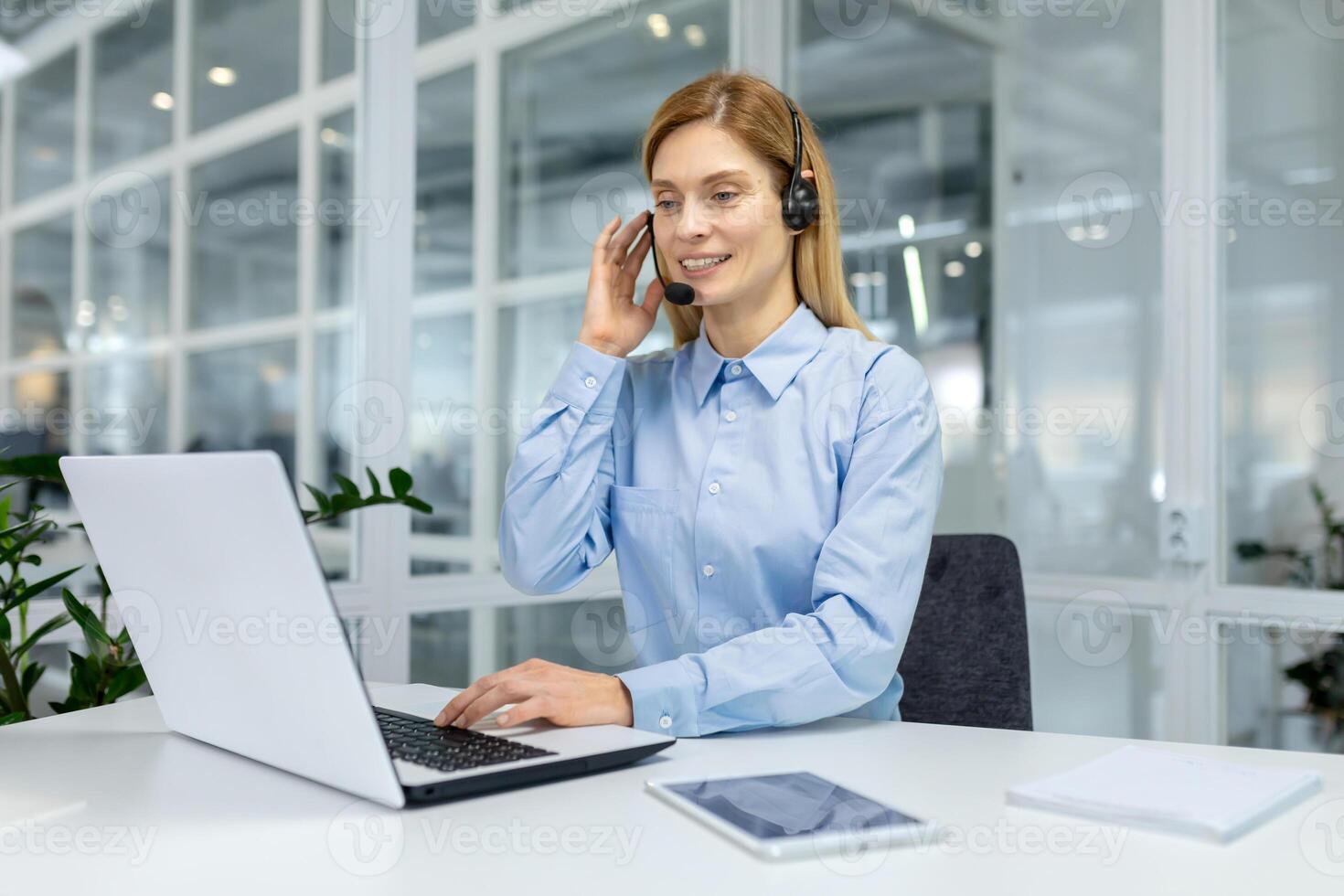 A focused professional woman in a blue shirt working with headphones at a bright office workspace with laptop. photo