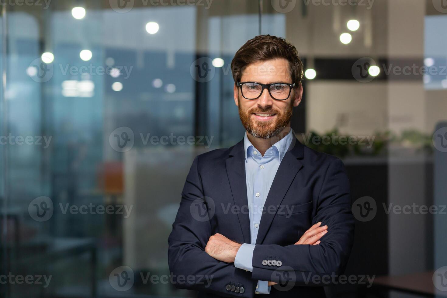 retrato de exitoso maduro jefe, mayor empresario en lentes y negocio traje mirando a cámara y sonriente, hombre con cruzado brazos trabajando dentro moderno oficina edificio. foto