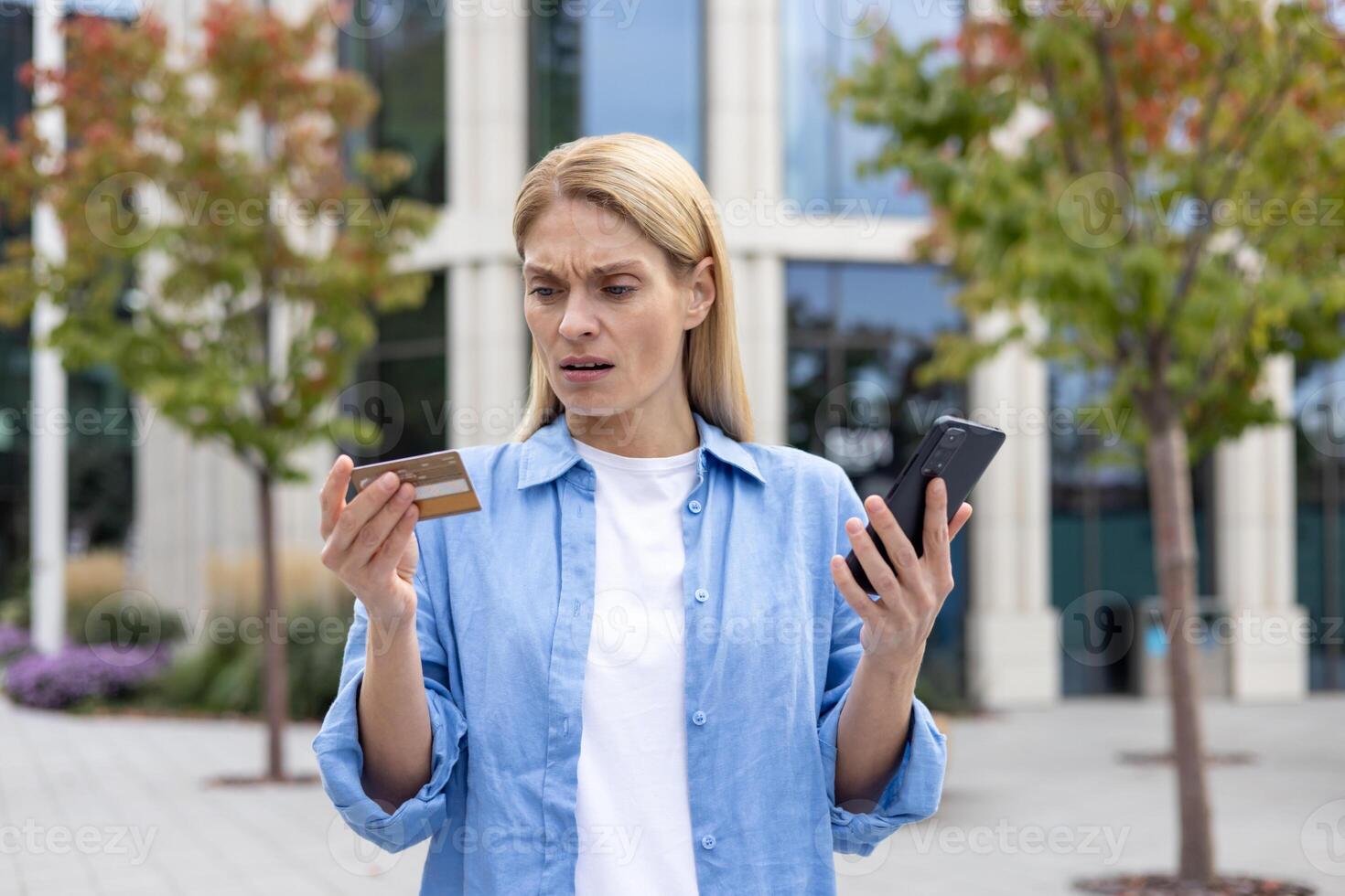 Deceived woman outside office building denied money transfer, mature female worker with phone and bank credit debit card standing, upset and unhappy blonde. photo