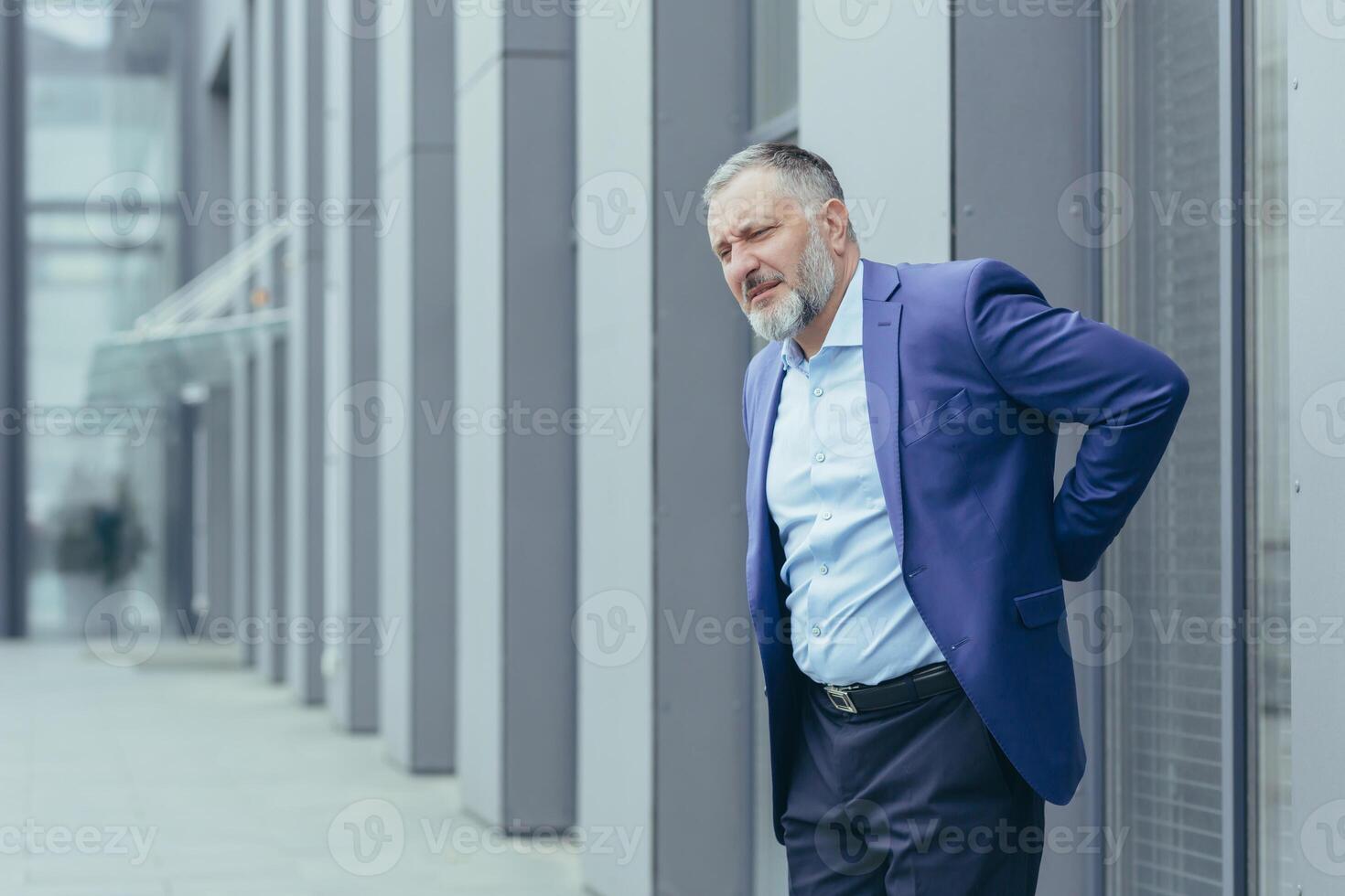 Senior male businessman suffering from back pain. Standing in a suit on the street near the office center, holding his hand behind his back, grimacing in pain, needs a break and medical help photo