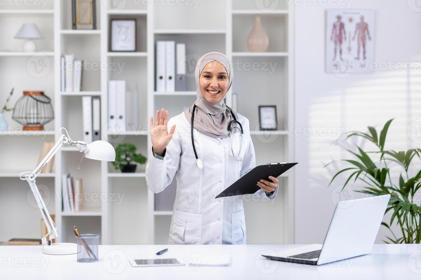 sonriente musulmán médico en hijab saludo con un ola mientras participación un portapapeles en un bien iluminado médico oficina. foto