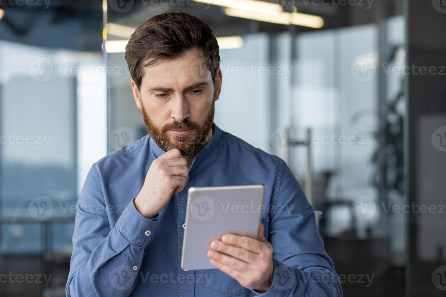 Close-up photo of a young man in a blue shirt who is seriously sitting at the desk in the office and thoughtfully looking at the screen ,of the tablet he is holding in his hands.