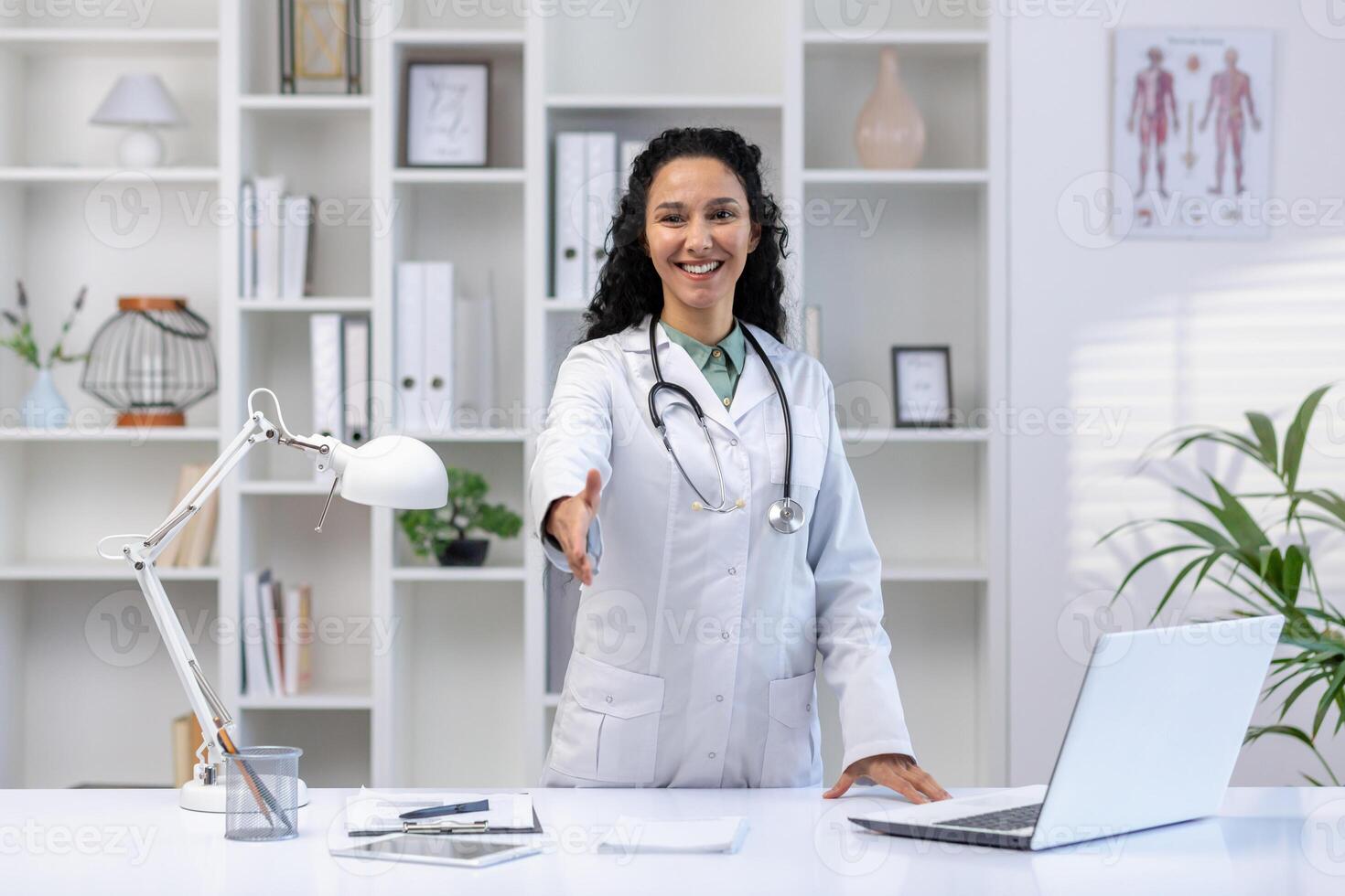 Young beautiful female doctor smiling and looking at camera standing inside clinic office, extending hand forward greeting gesture, Hispanic woman in white medical coat female worker. photo