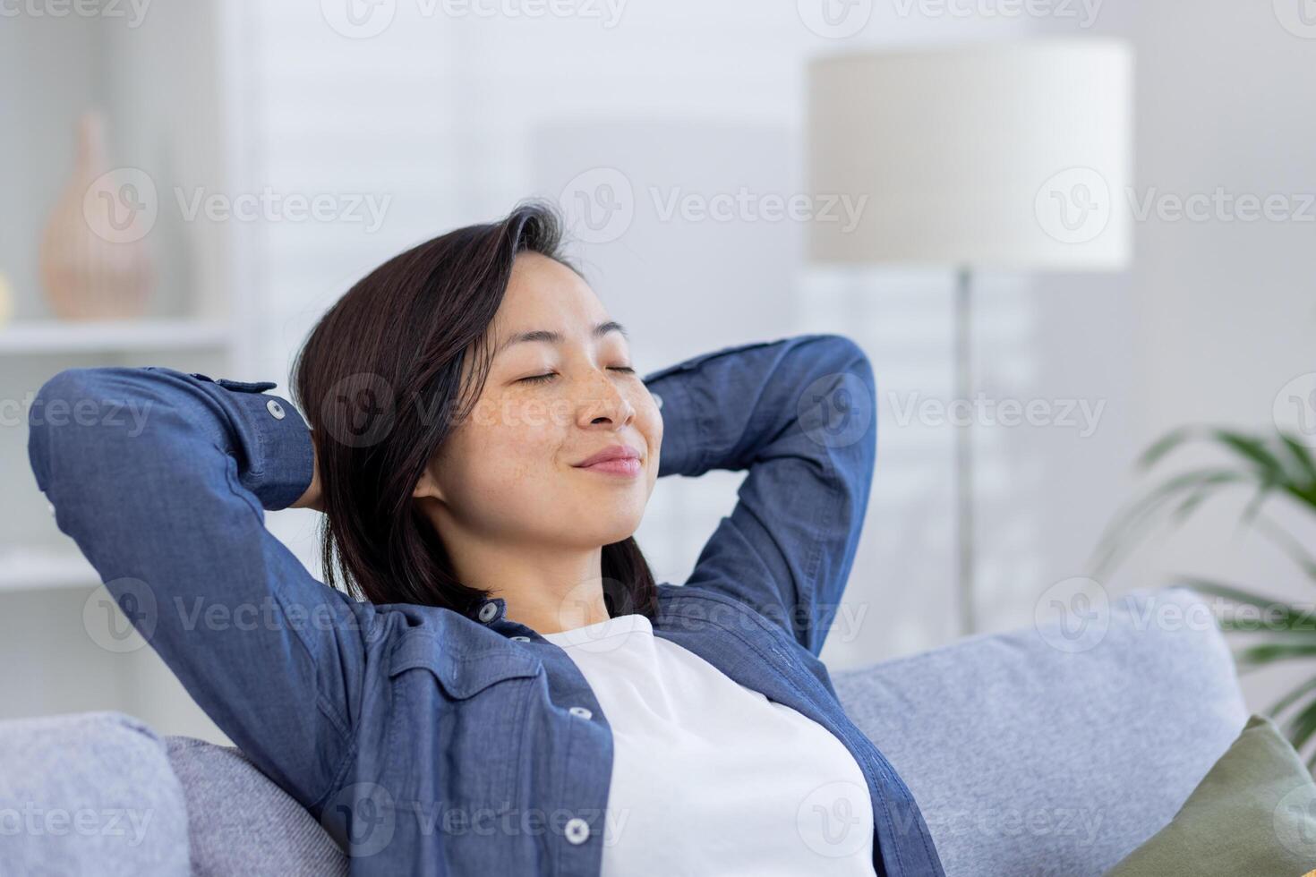 Young beautiful Asian woman relaxing at home close-up on sofa in living room, hands behind head with eyes closed dreaming and visualizing future results achievement and plans. photo