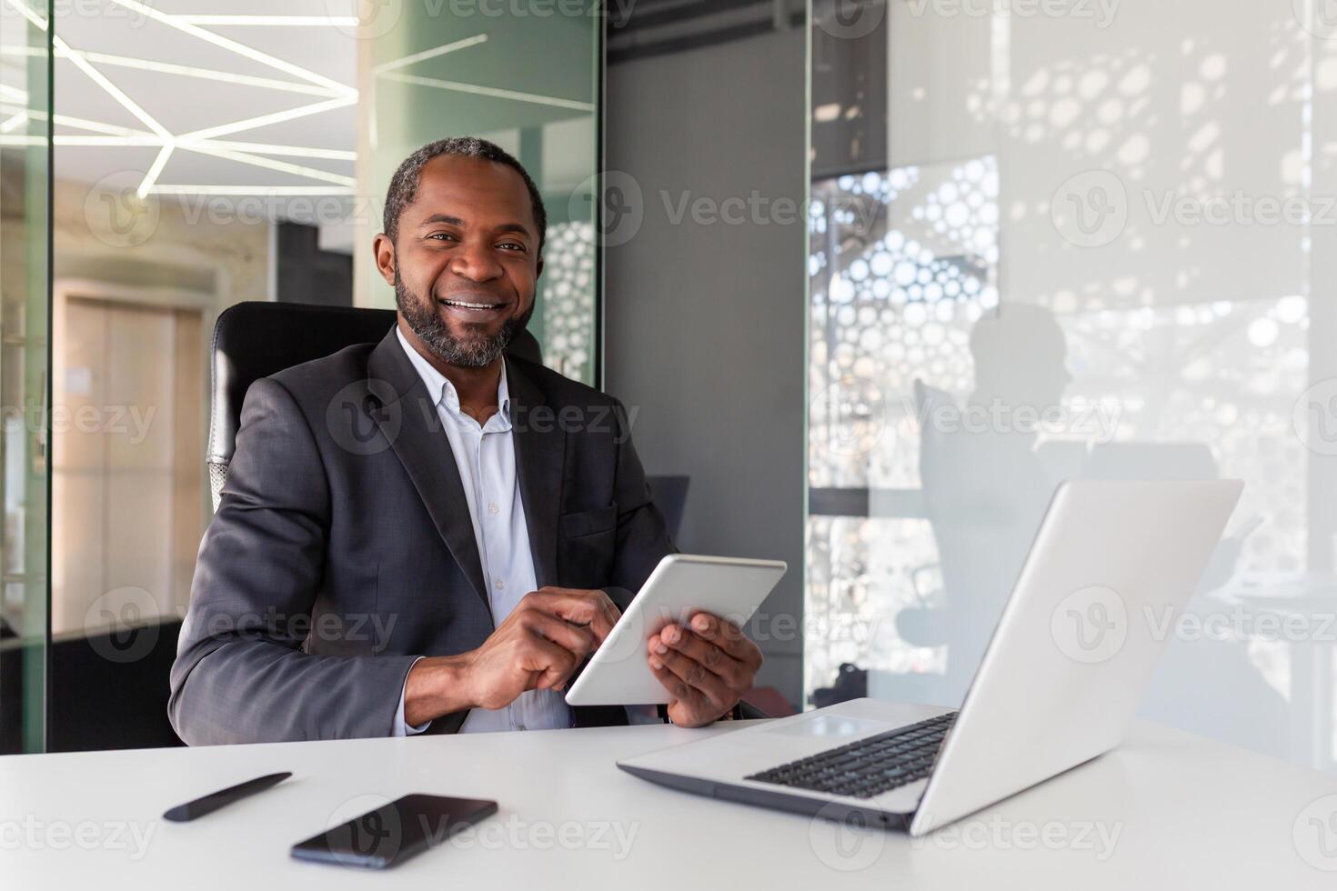 Portrait of successful African American businessman inside office, boss with tablet computer smiling and looking at camera, man at workplace happy with financial results and achievements. photo