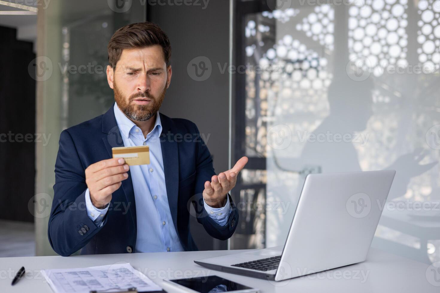 Upset young man businessman sitting at a desk in the office in front of a laptop and looking disappointedly at the credit card. Problem with account and compromised security password. photo