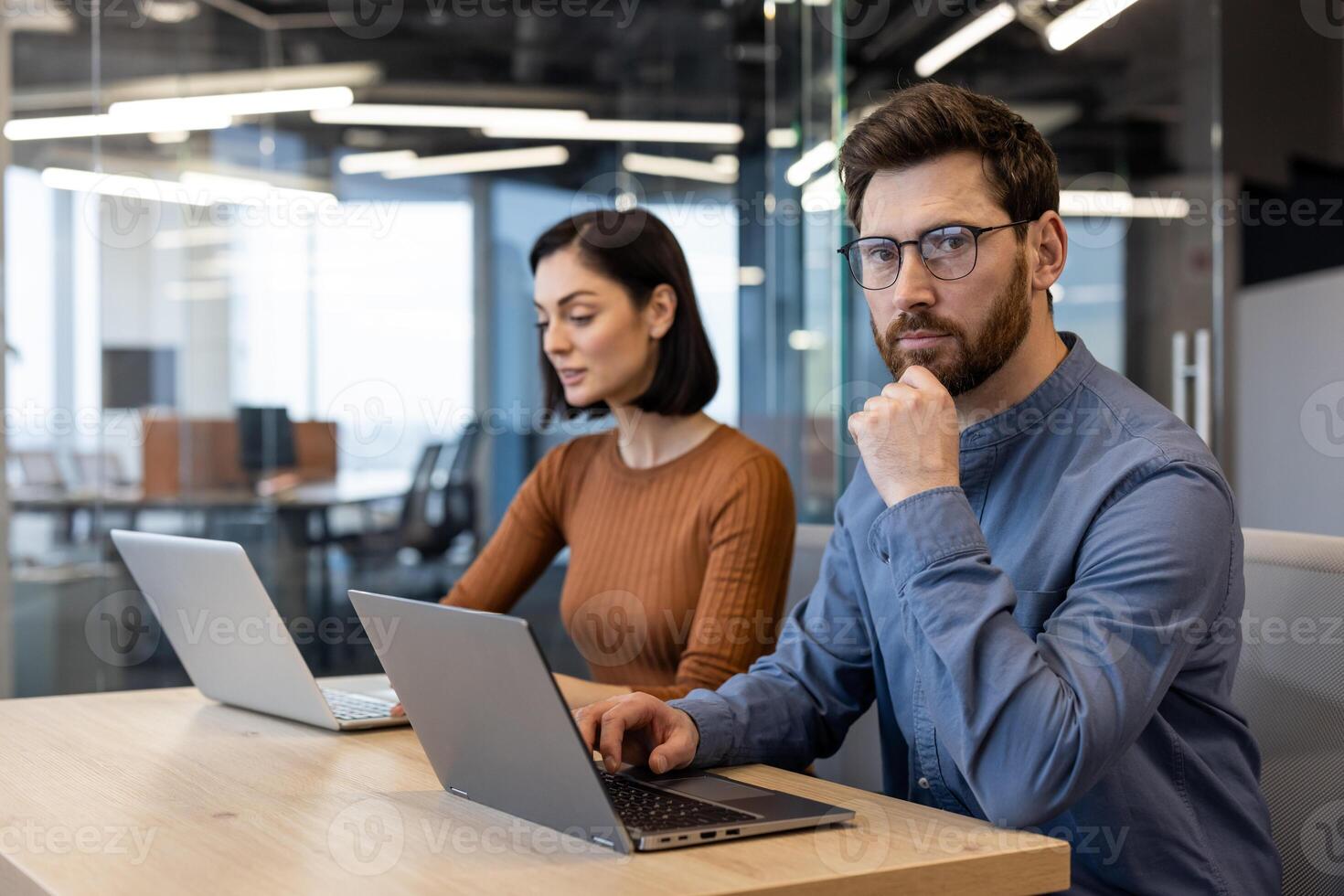 Pensive unshaved guy in denim shirt resting head on hand while looking at camera next to female colleague. Two digital designers developing new creative strategy for social media for marketing team. photo