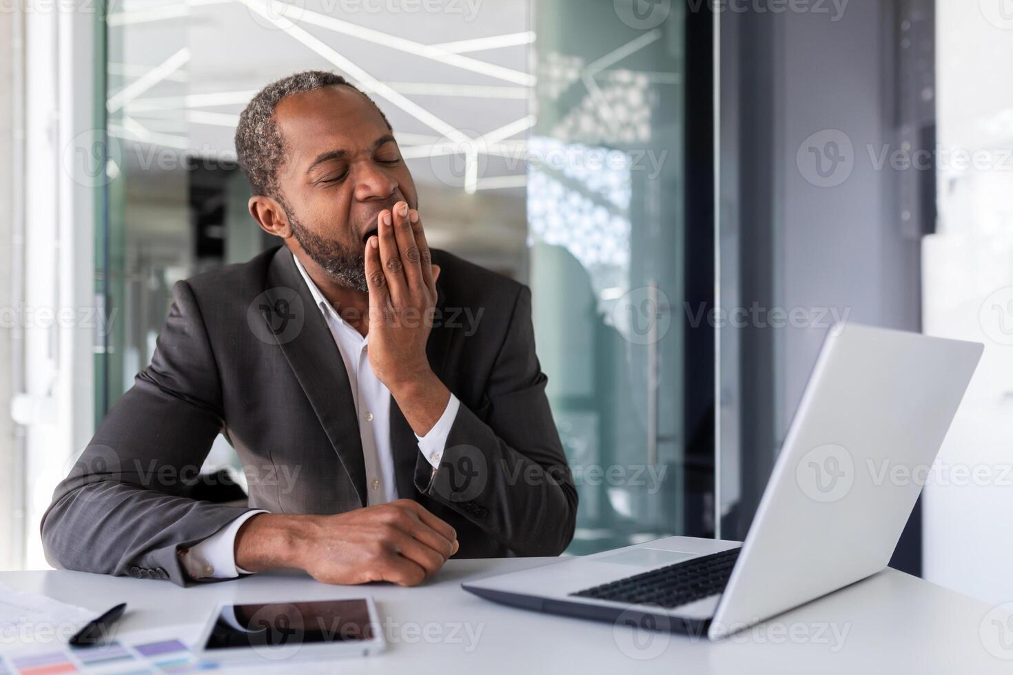 Tired businessman misses boring work and everyday, african american mature experienced man in business suit yawns, at workplace inside office. photo