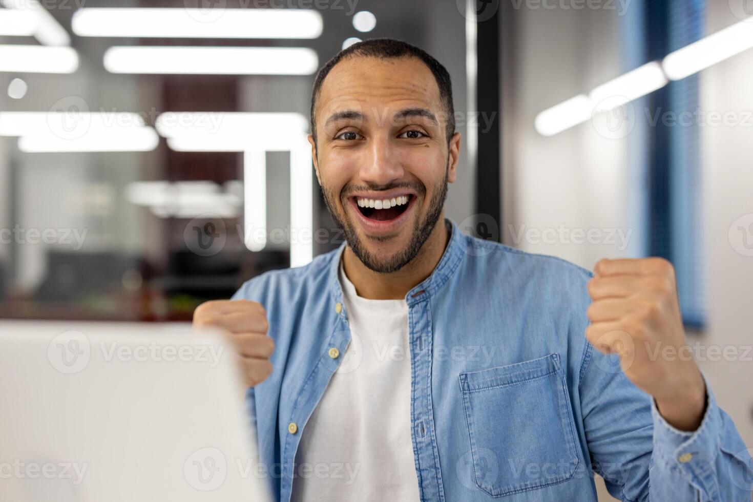 Close-up portrait of a happy young hispanic man sitting in a modern office, in front of a laptop and looking at the camera with a victory gesture. photo