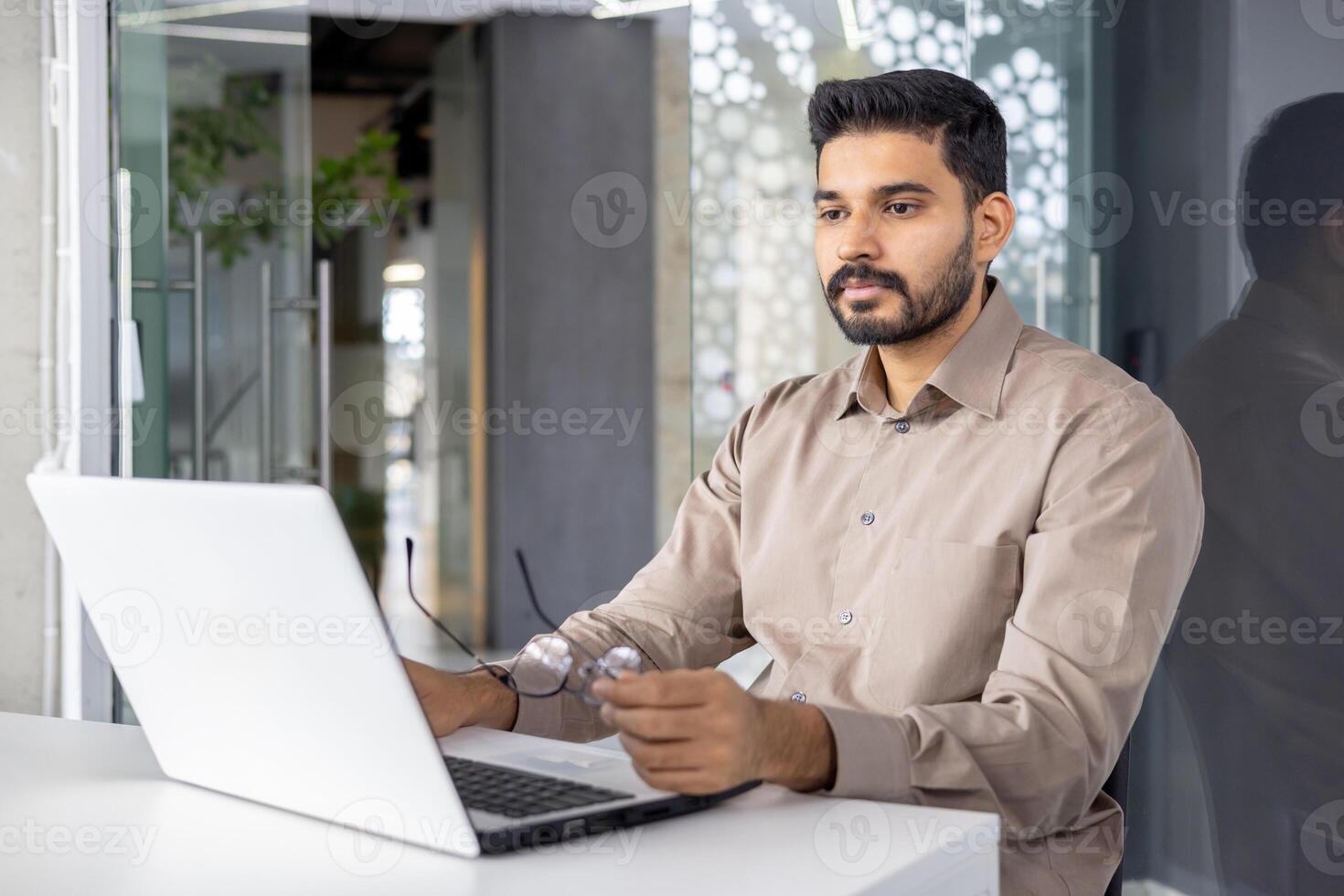 A focused professional man sits at a desk in a contemporary office, working intently on his laptop. His thoughtful expression and business casual attire reflect a typical workday environment. photo