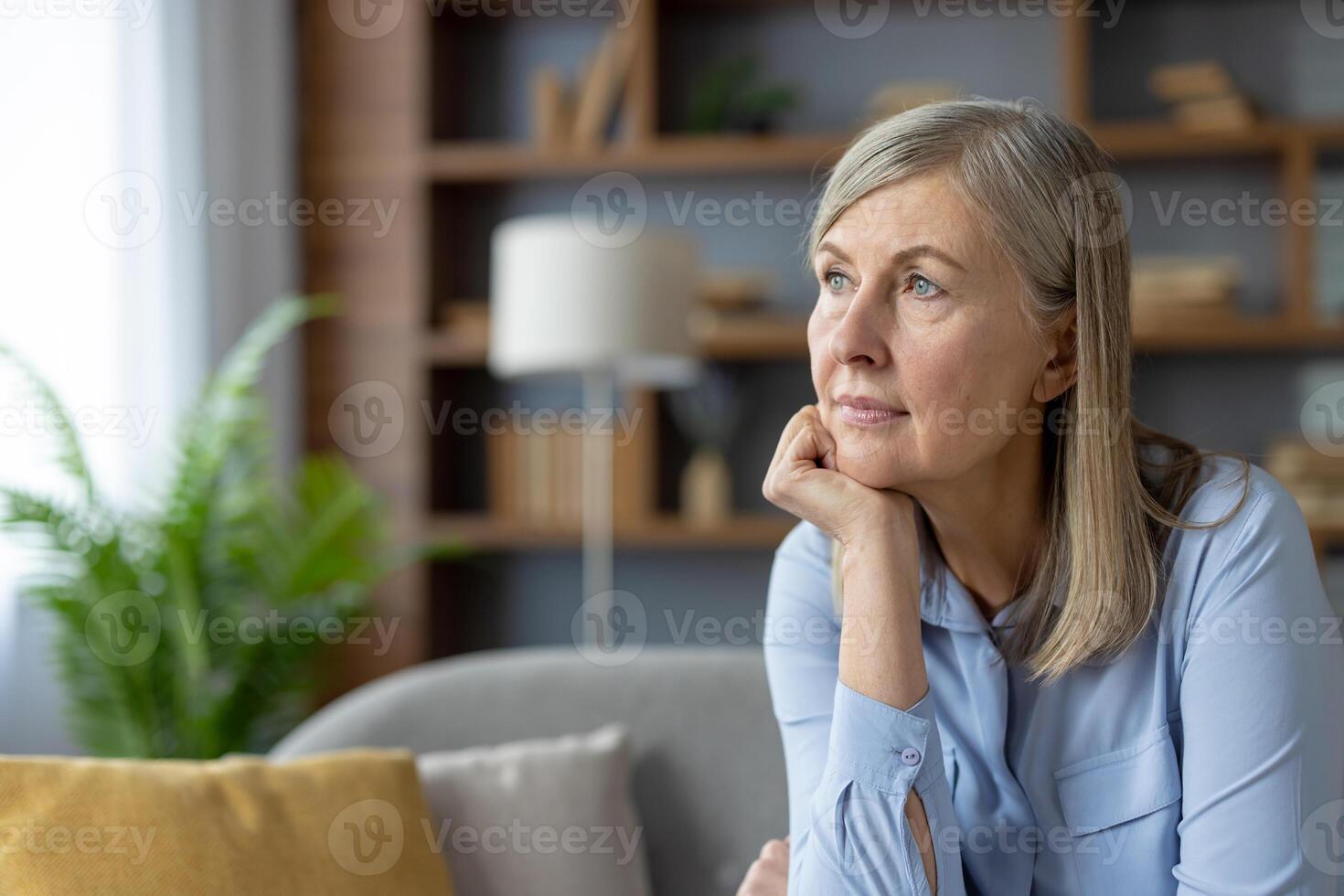 un sereno maduro mujer en un azul camisa miradas pensativamente mientras sentado en un bien iluminado vivo habitación. el pacífico atmósfera Destacar su pensativo humor. foto