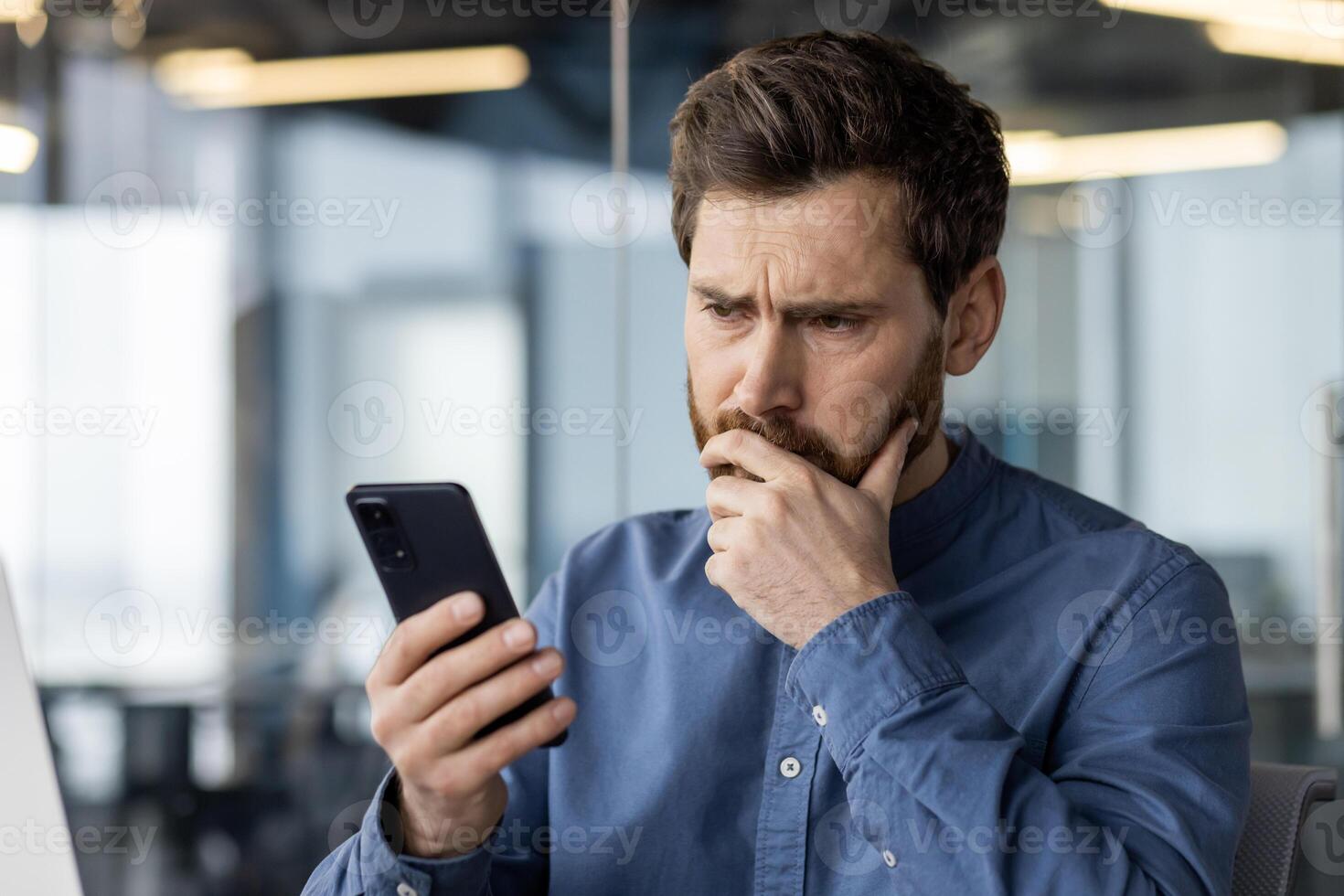 A focused male adult with a beard looks at his smartphone with a worried expression, possibly dealing with urgent business matters or bad news. photo