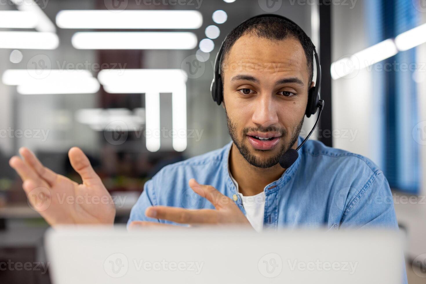 Close-up photo of a smiling young hispanic man in a headset sitting in the office in front of a laptop and talking on a call ,while gesturing with his hands.