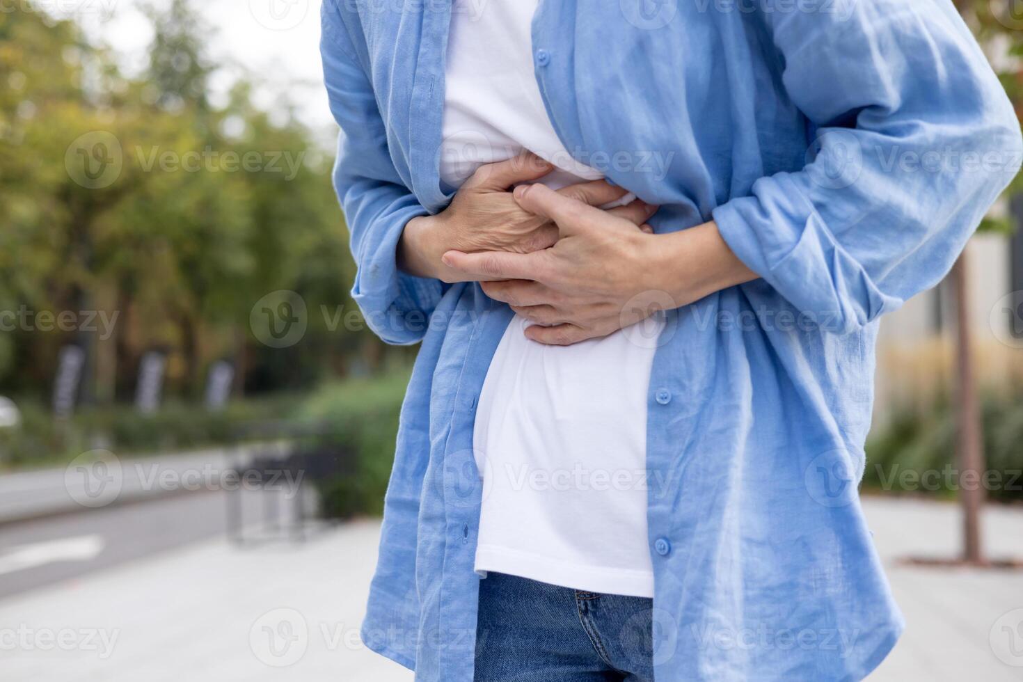 Unrecognizable person in casual clothes grabbing stomach area with hands while standing in public zone. Sick human in blue linen shirt suffering from digestion problems after eating street food on go. photo