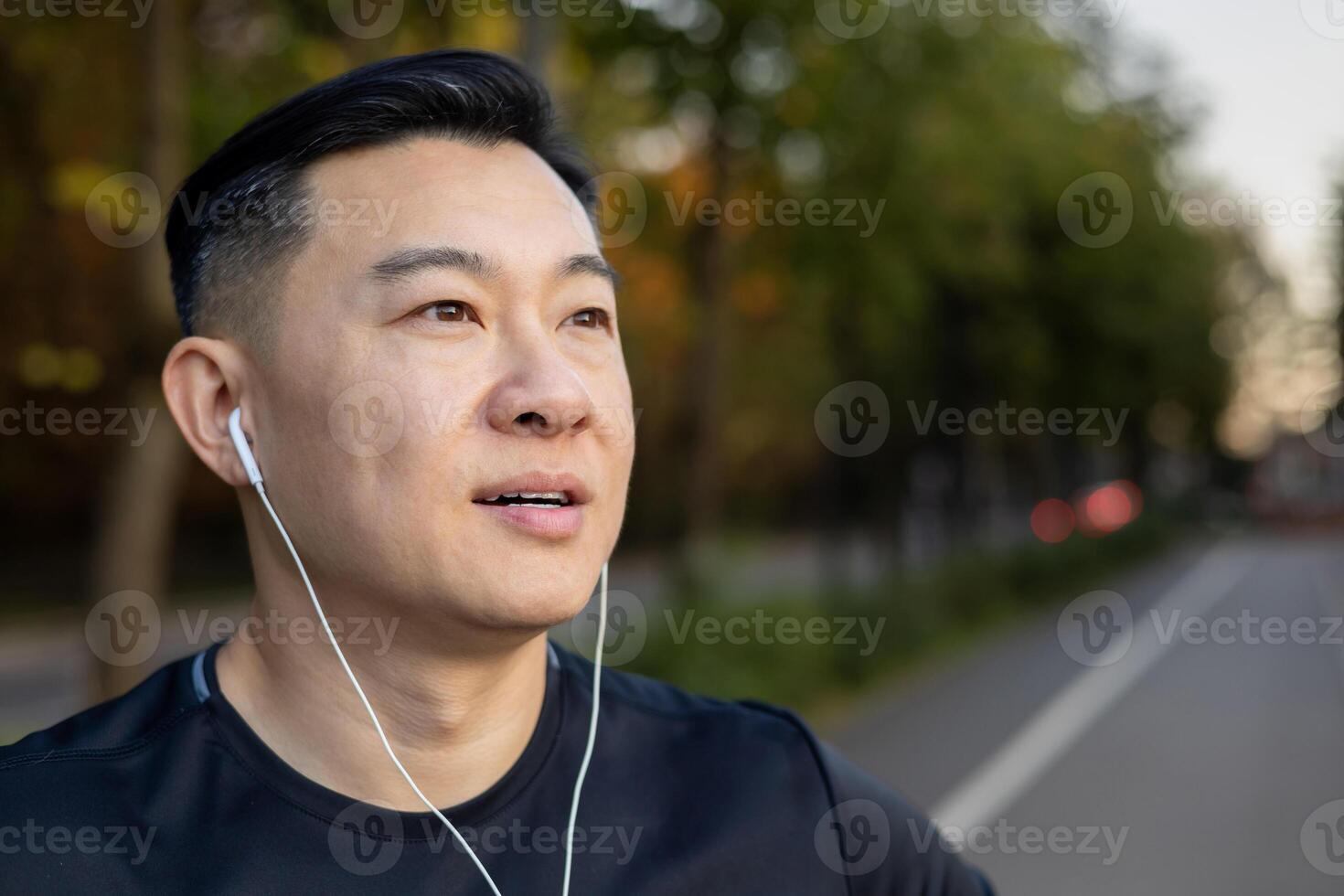 un de cerca foto de un joven grave hombre asiático masculino deportista en pie en el la carretera en el medio de el calle en auriculares, escuchando a música, descansando, mirando a el lado.