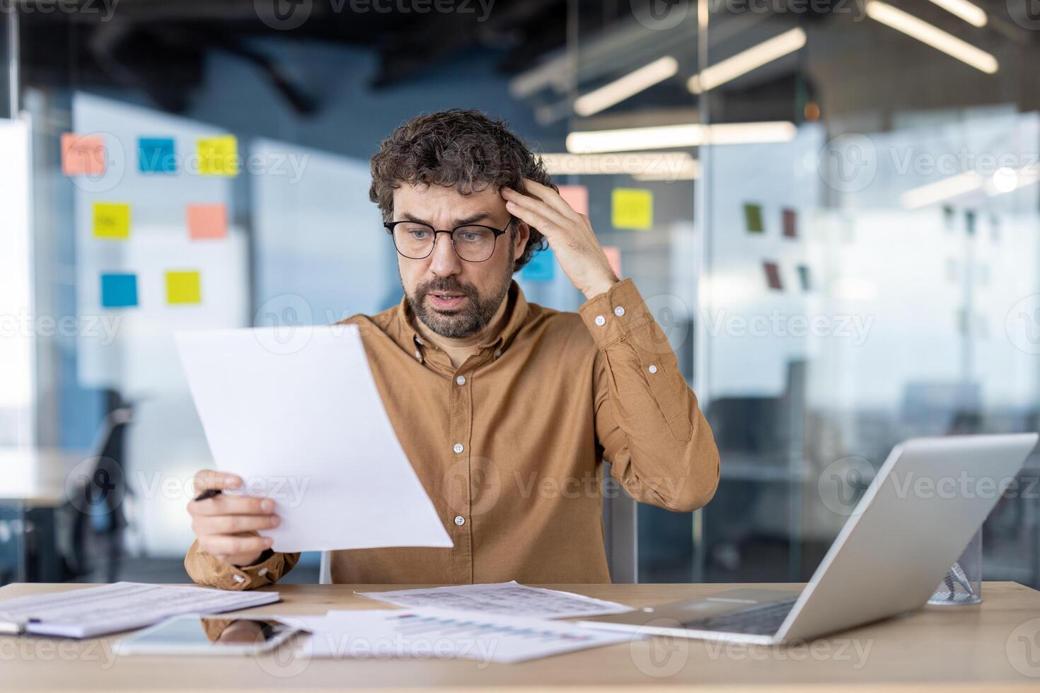 A perplexed mature adult man in glasses scratches his head while looking at papers with a laptop open in an office setting. photo