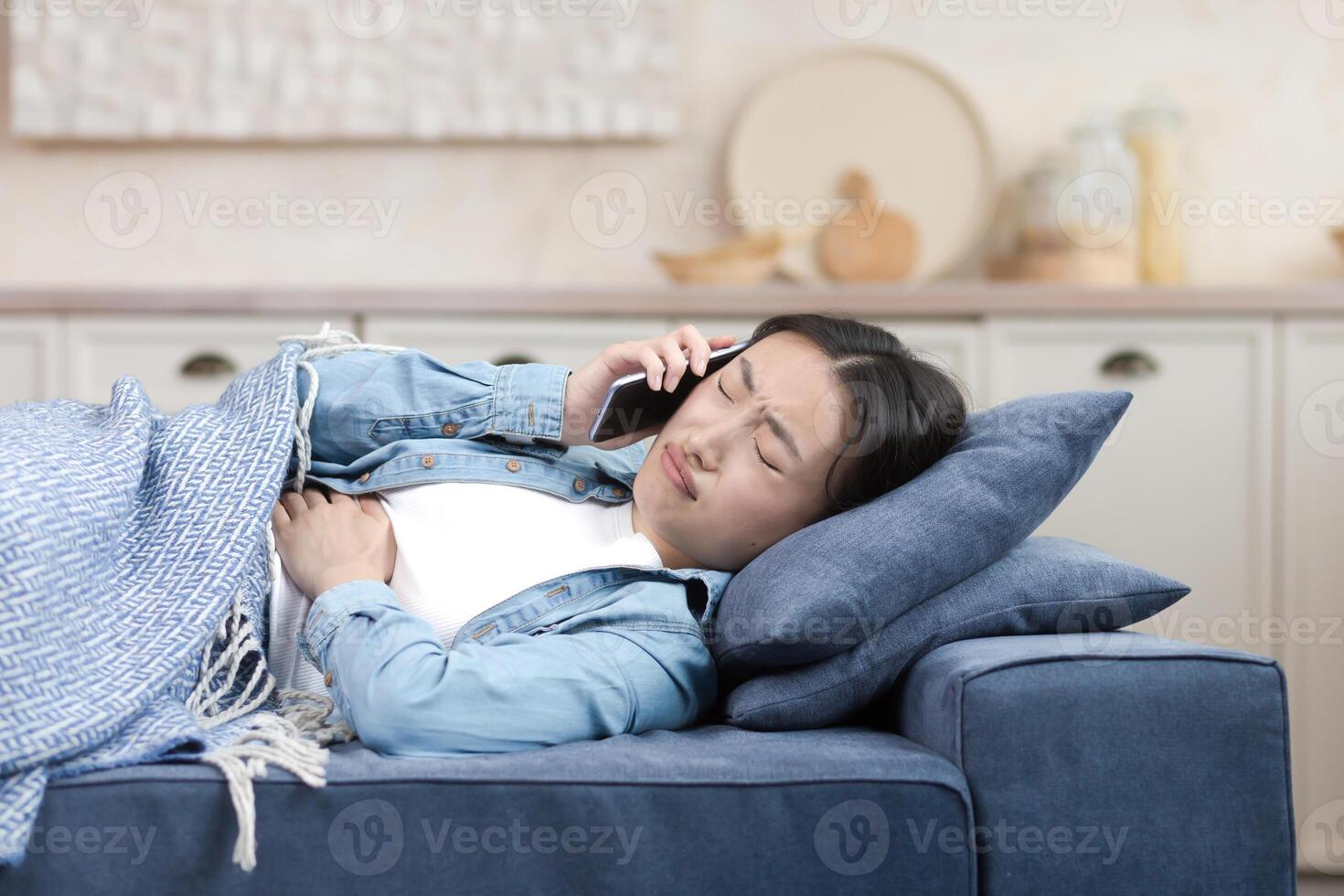 A sick Asian woman at home is lying on the sofa under a blanket, she has a severe stomach ache, the woman calls the doctor and asks for help photo