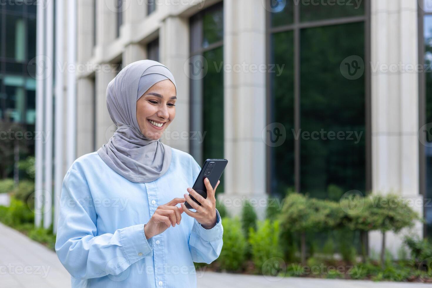 Young beautiful business woman in hijab walks in the city outside the office building, Muslim woman uses an application on the phone, browses online internet pages, smiles contentedly. photo