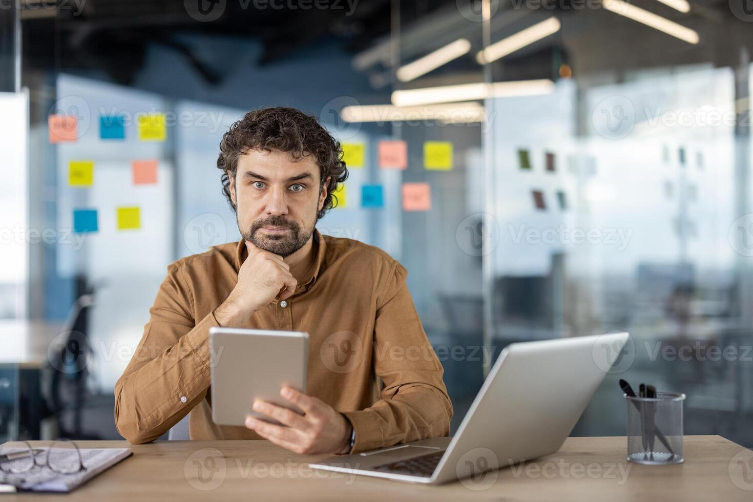 Focused professional using a laptop and a tablet at a wooden desk in a modern office, with sticky notes in the background. photo
