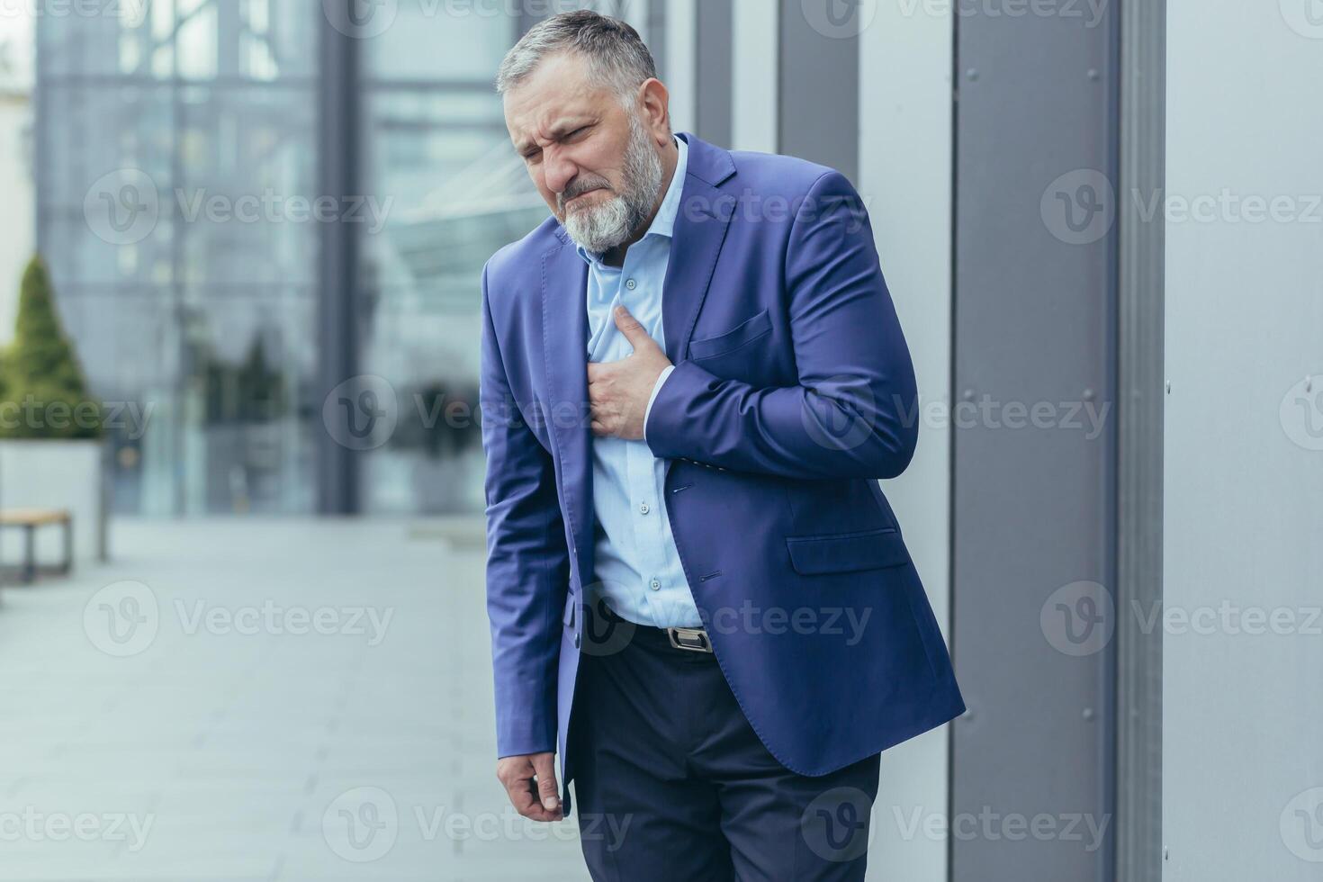 Pre-infarction condition. A senior man in a suit stands on the street near an office center holding his heart. He feels a sharp pain in his chest. He needs urgent medical help. photo