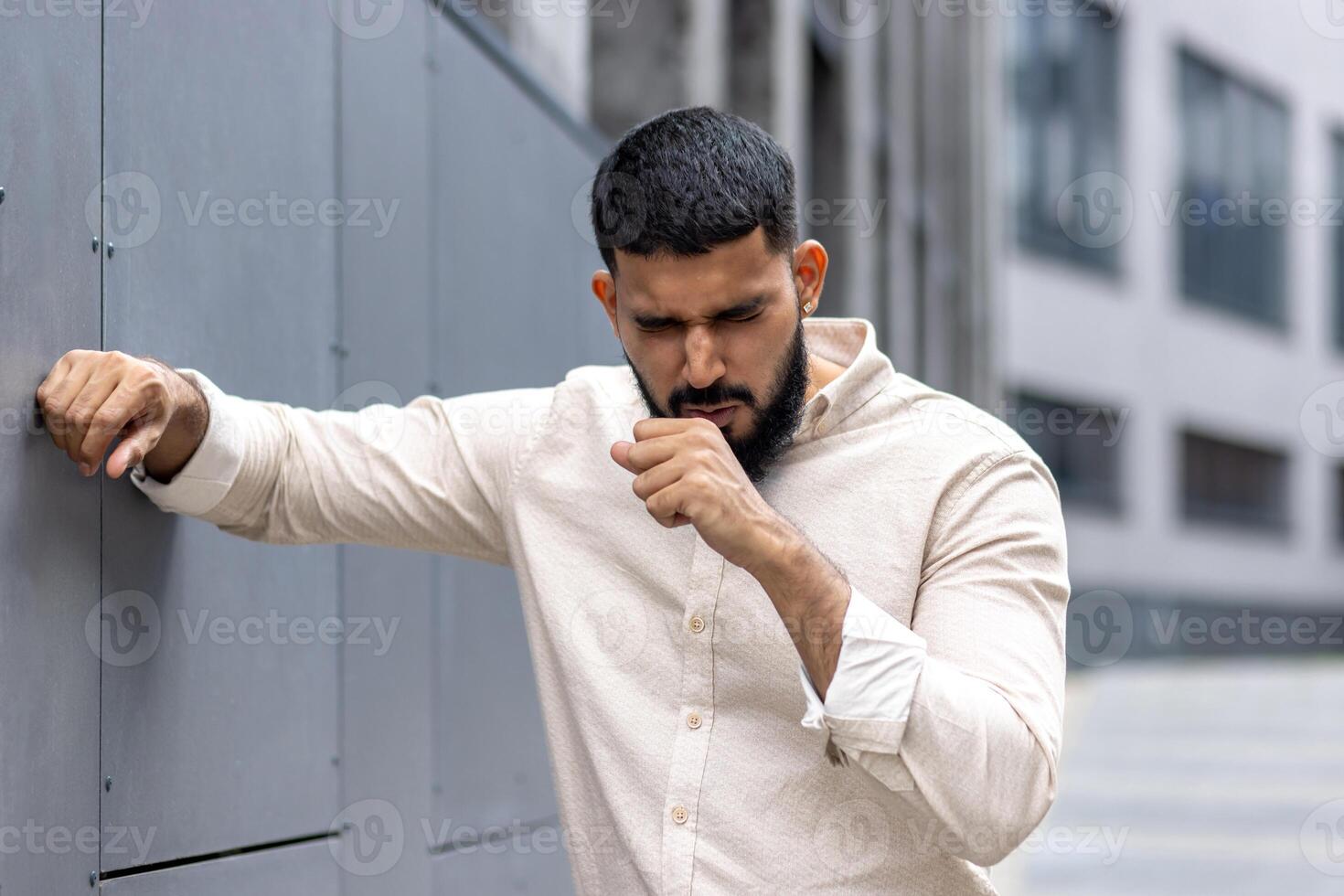 Cough attack. A young Indian man is standing outside the building, holding on to the wall with his hand, covering his mouth with his hand and coughing, breathing hard. Close-up photo