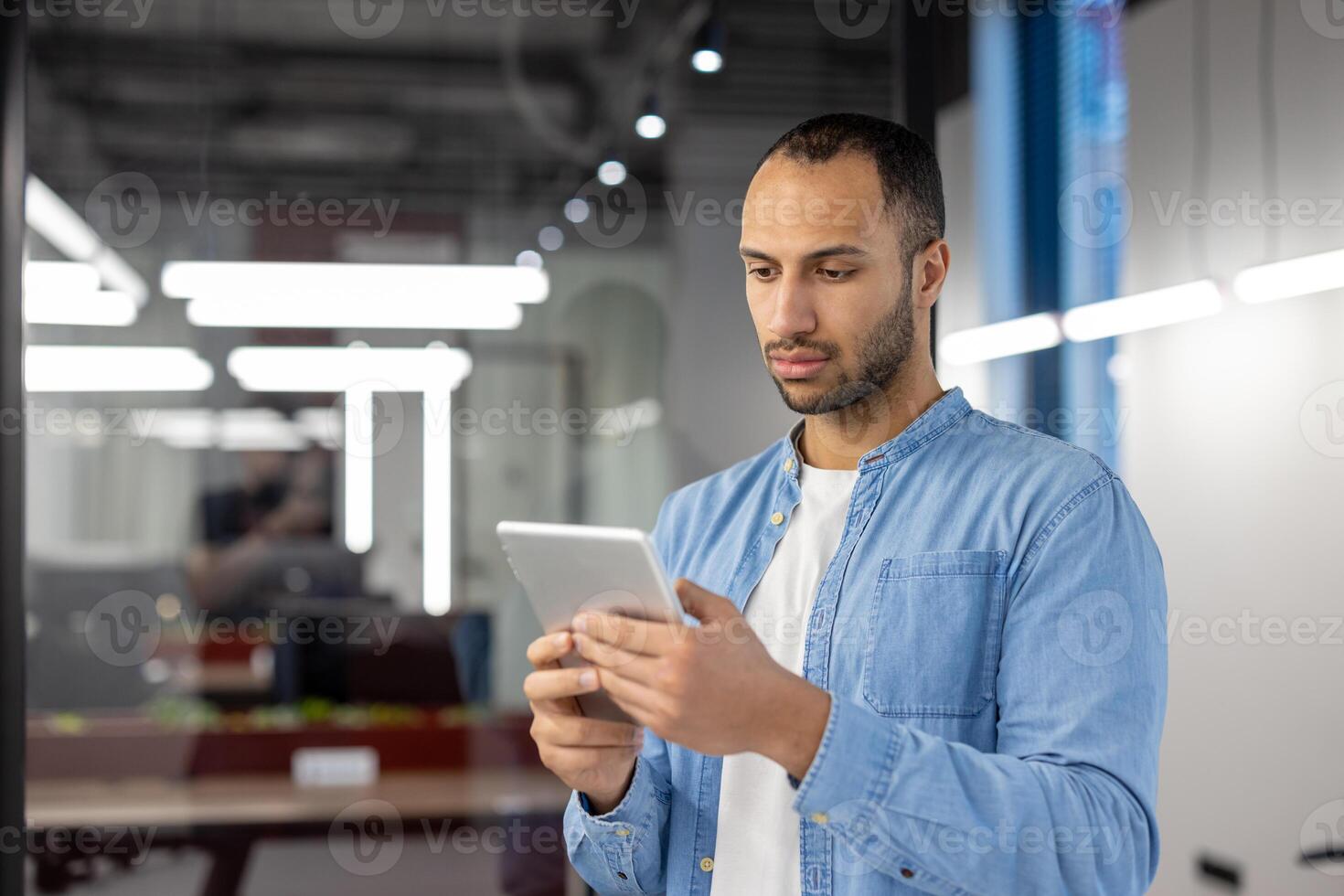 Focused Indian man in casual business attire using a digital tablet inside a contemporary office. Concept of technology at work. photo
