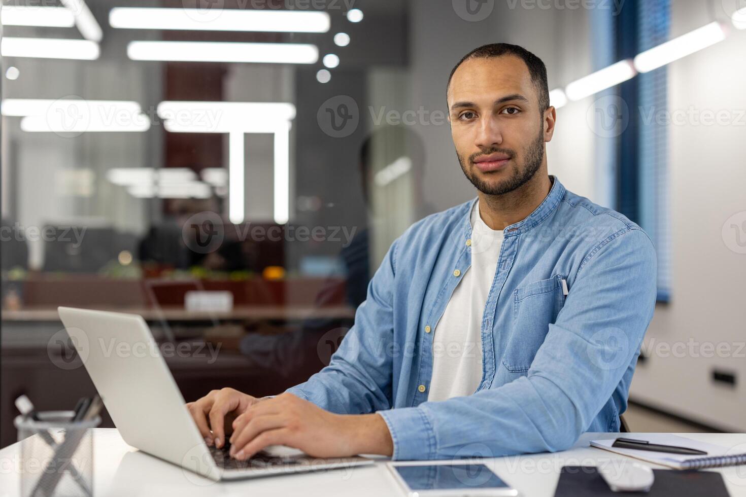Portrait of a young confident Muslim man working and studying in the office, sitting at a desk with a laptop in casual clothes and looking confidently at the camera. photo