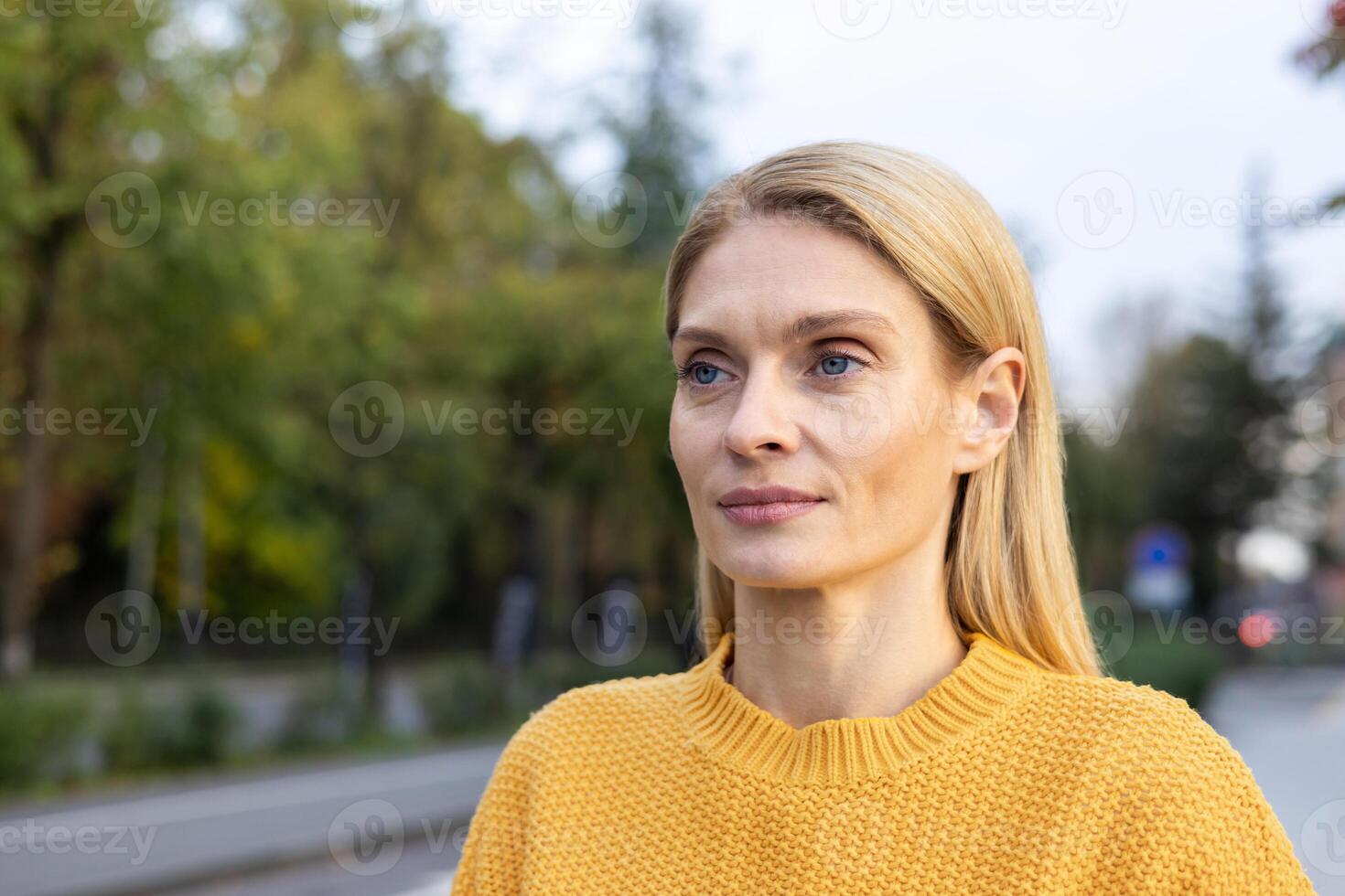 Close-up of a mature Caucasian woman in a yellow sweater, looking contemplative as she enjoys a peaceful autumn day outdoors. The background is softly blurred with hints of greenery. photo