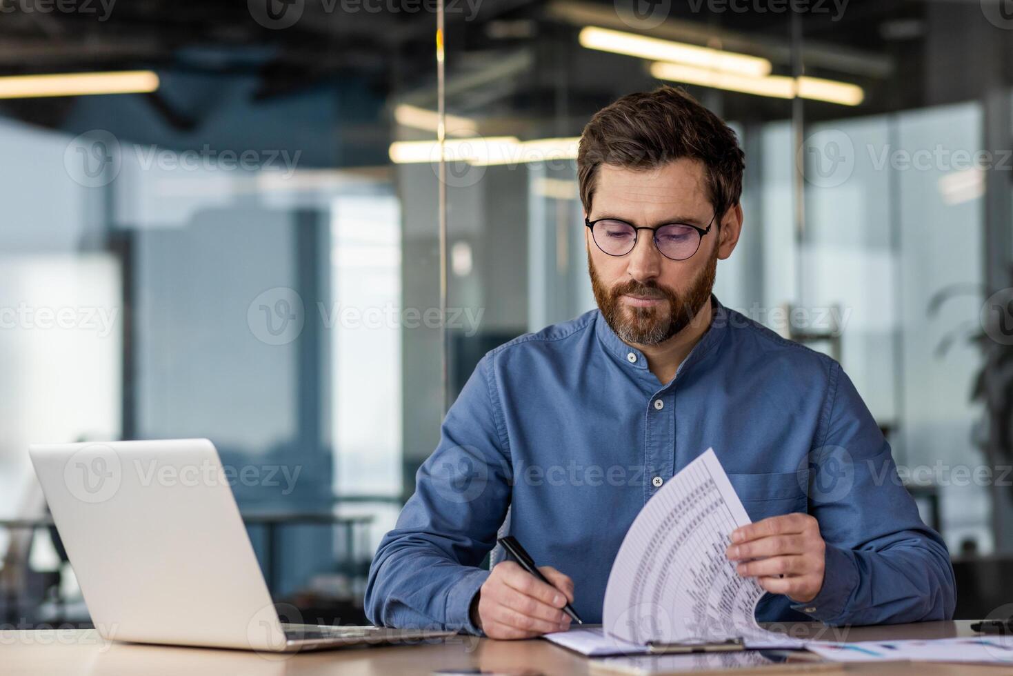 A young serious business man is sitting in the office at a desk with a laptop and is focused on working with documents. Writes and checks data and invoices. photo