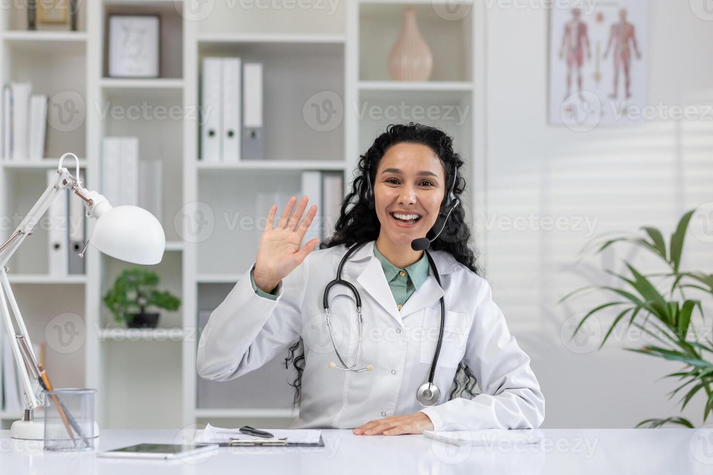 A cheerful female physician engaging in a call, waving to the camera from her office, equipped with medical literature and plants. photo