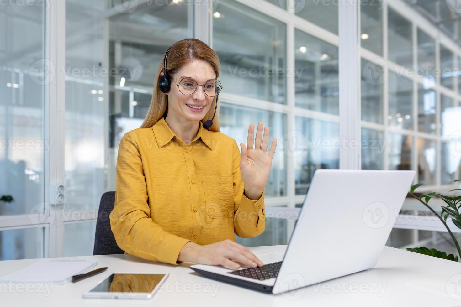 Office worker works remotely, online customer support service, help service, woman uses headset and laptop for online consultation and call, works inside office, waves greeting gesture. photo