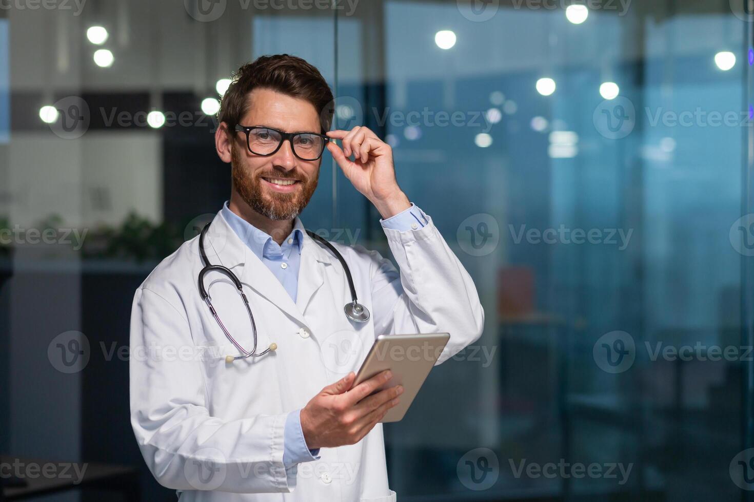 Portrait of a young handsome male doctor. He stands in the office in a white coat and with a stethoscope. He holds a tablet in his hands. He looks at the camera, adjusts his glasses, smiles. photo