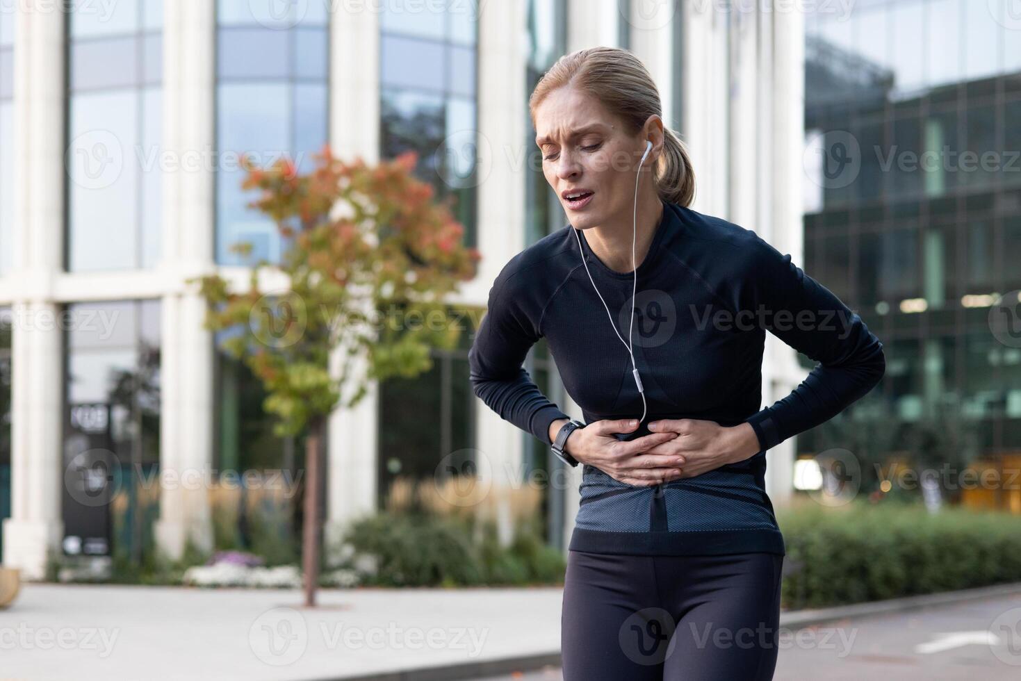 Tired overwhelmed woman holding hands on stomach senior mature runner during jogging and fitness class has severe stomach pain bent over massaging muscle. photo