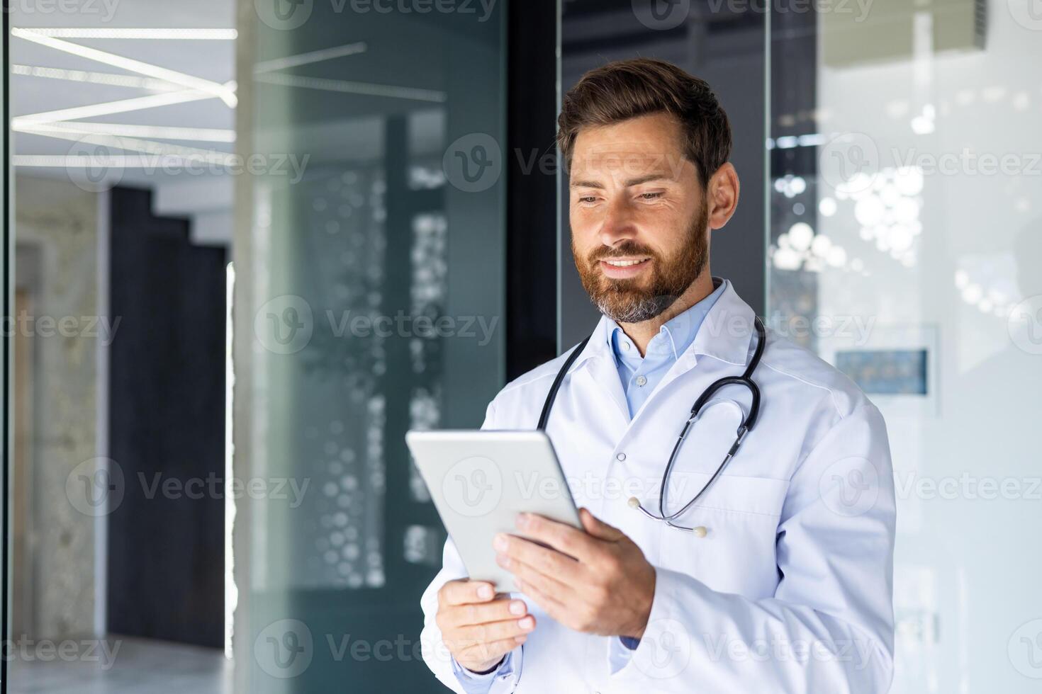 Close-up photo of a smiling young male doctor standing in a clinic in a white coat and using a tablet.