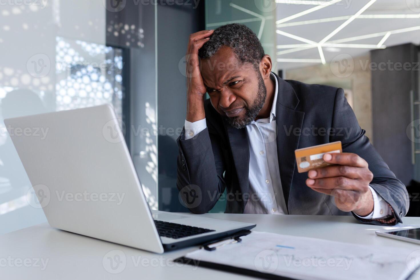 trastornado y preocupado empresario a lugar de trabajo, hombre engañado recibido en línea dinero transferir error, africano americano hombre infeliz sentado con ordenador portátil dentro oficina participación banco crédito tarjeta. foto