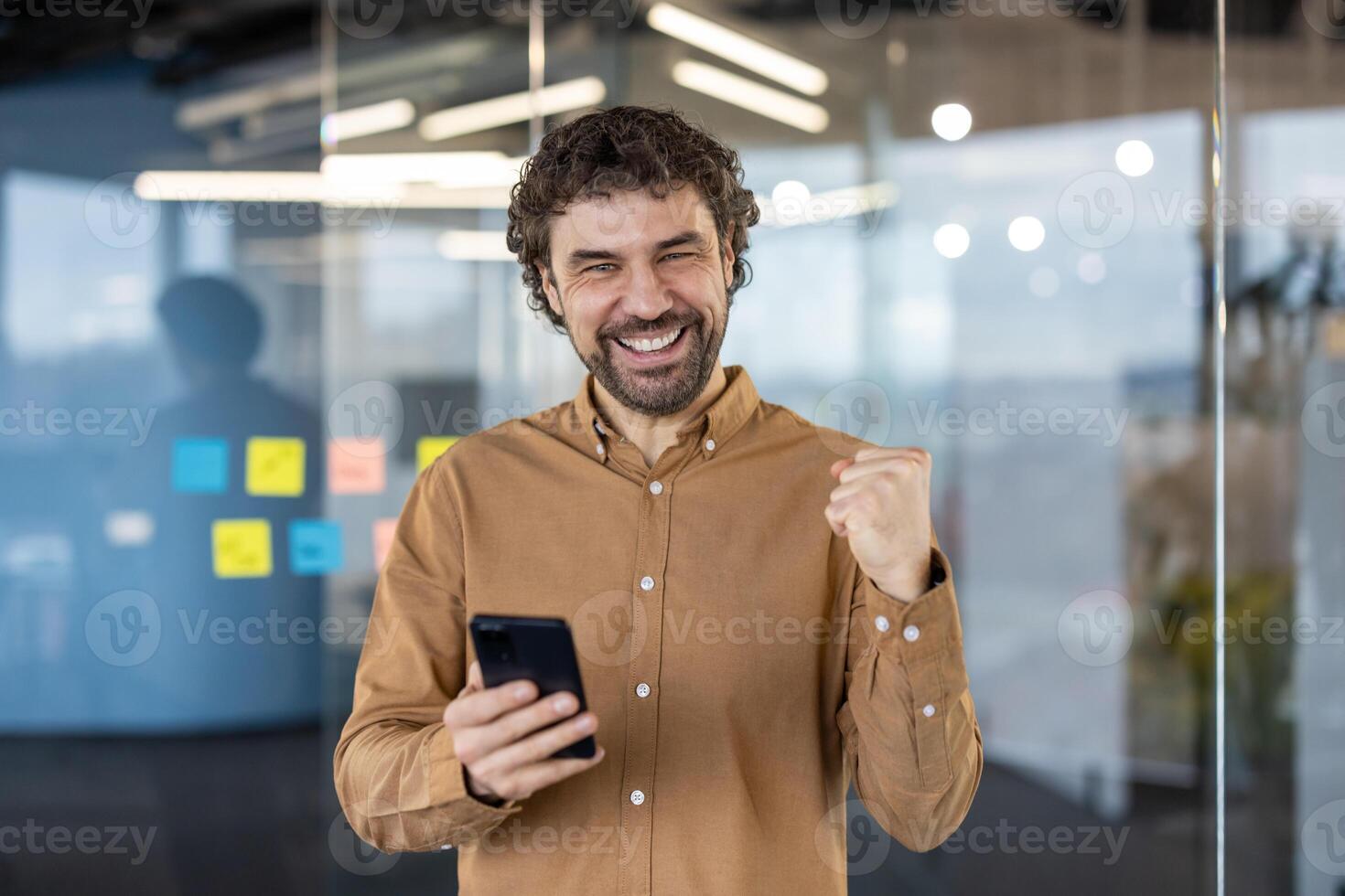 un alegre empresario en un casual camisa celebra un éxito con un puño bomba mientras participación un teléfono inteligente en un moderno oficina ambiente. foto