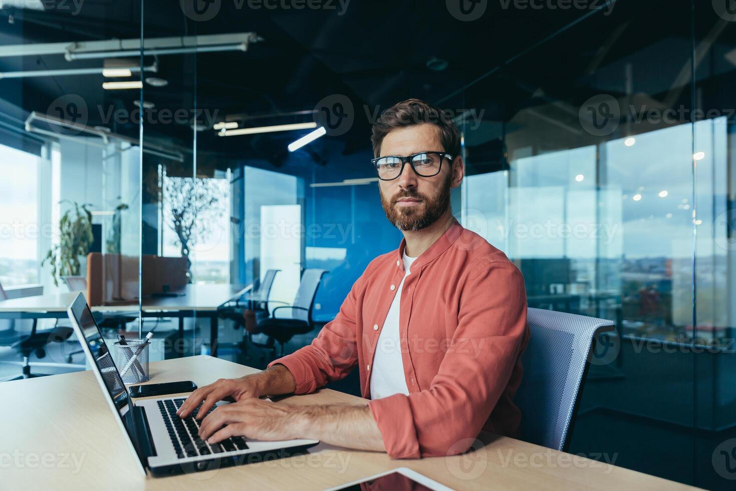 retrato de un exitoso programador dentro el oficina, un hombre con un barba, lentes y un rojo camisa mira a el cámara, un desarrollador ingeniero usos un ordenador portátil a escribir código en un programación estructura. foto