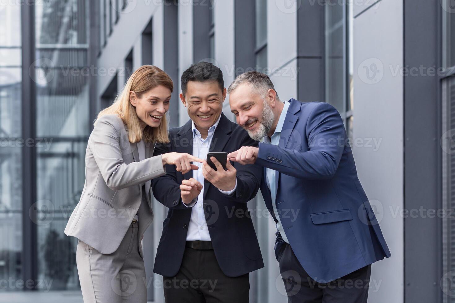 Diverse business group, three male and female workers outside office building cheerfully smiling and happy watching on phone photo