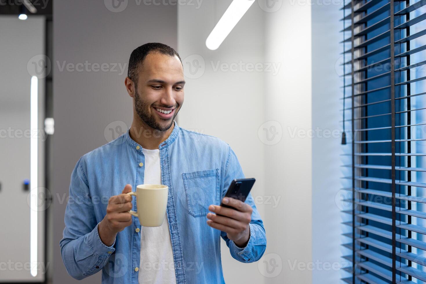 Smiling young arab man working in office, standing by window on work break, holding cup with drink and using mobile phone. photo