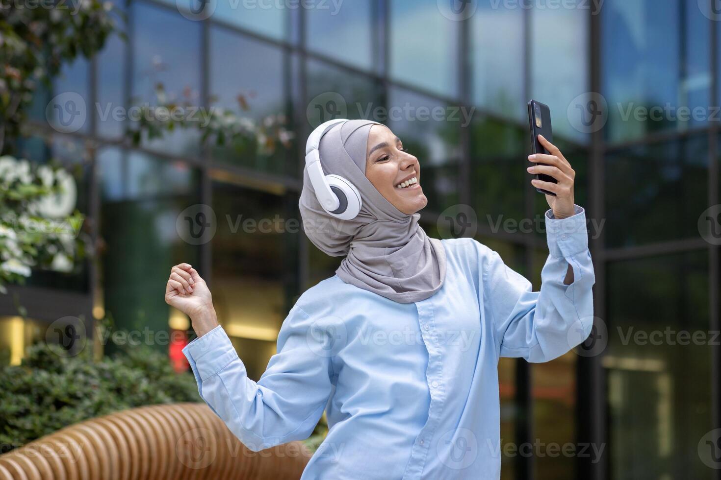 A modern Muslim woman in a hijab is capturing a selfie with her smartphone outside a reflective glass office building. photo