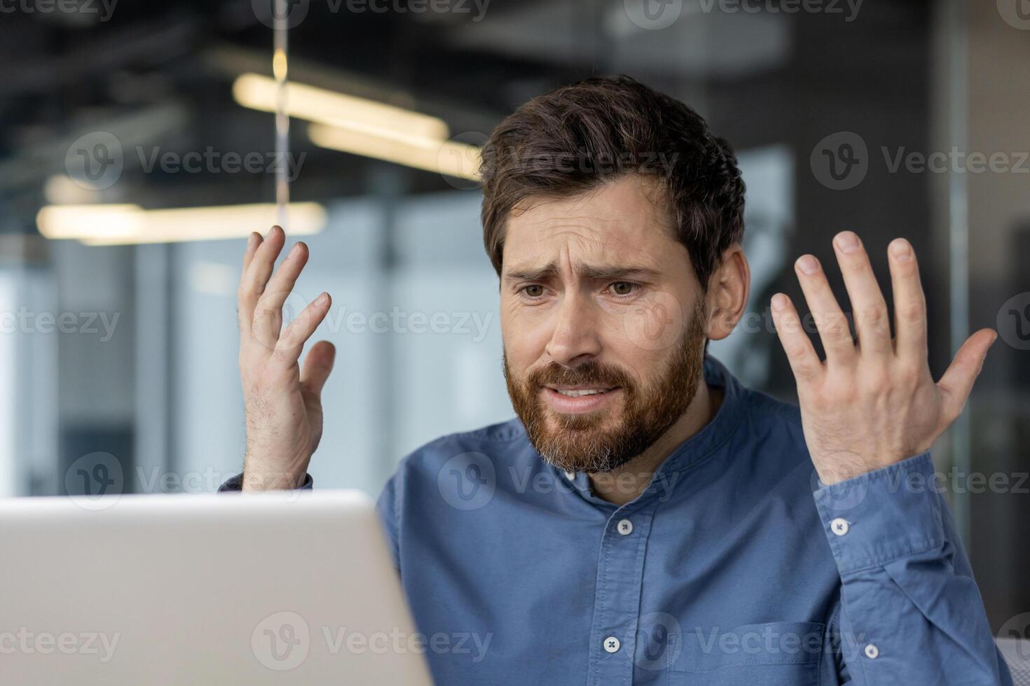 A confident young man sits in a modern office and looks disappointedly at the laptop screen, spreading his hands. Close-up photo. photo